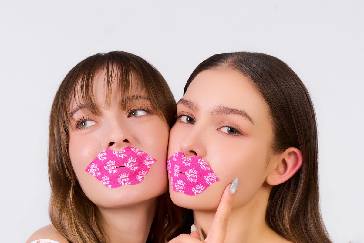 Two friends cheek to cheek wearing hot-pink Queen Tape Hydrating Collagen Mouth Tape, one giving a playful side-eye